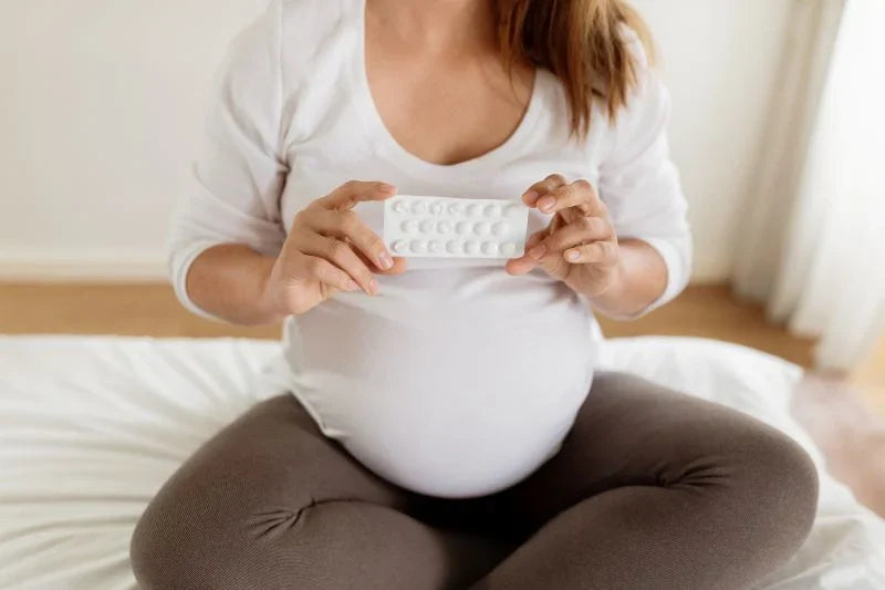 A pregnant woman holding a pack of prenatal supplements, sitting on the bed and preparing to take her daily vitamins for a healthy pregnancy